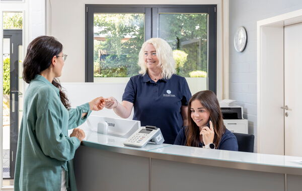 Eine Patientin übergibt einer Praxismitarbeiterin eine Karte am Anmeldetresen, eine andere Mitarbeiterin telefoniert.