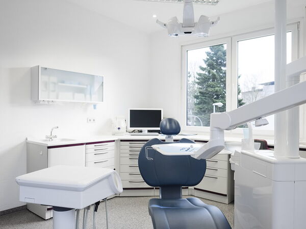 Ein Behandlungszimmer der Zahnarztpraxis Dr. Schlotthauer mit einem blauen Stuhl, weißen Geräten sowie im Hintergrund ein Bildschirm auf dem Tisch.