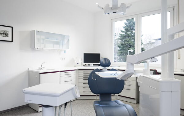 Ein Behandlungszimmer der Zahnarztpraxis Dr. Schlotthauer mit einem blauen Stuhl, weißen Geräten sowie im Hintergrund ein Bildschirm auf dem Tisch.