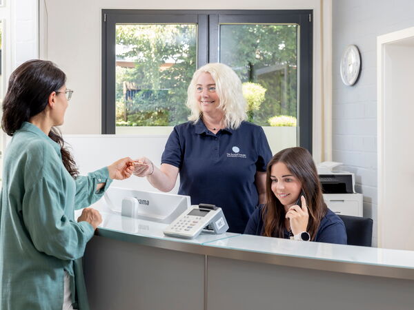 Eine Patientin übergibt einer Praxismitarbeiterin eine Karte am Anmeldetresen, eine andere Mitarbeiterin telefoniert.