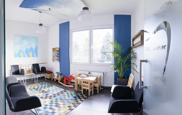 Das helle, moderne Wartezimmer der Zahnarztpraxis Dr. Schlotthauer mit Stühlen, einem Kinderspielbereich und einer grünen Pflanze.