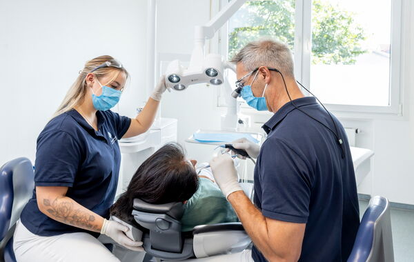 Eine Patientin liegt auf der Behandlungsliege und wird von Dr. Schlotthauer an den Zähnen untersucht, eine Mitarbeiterin assistiert.