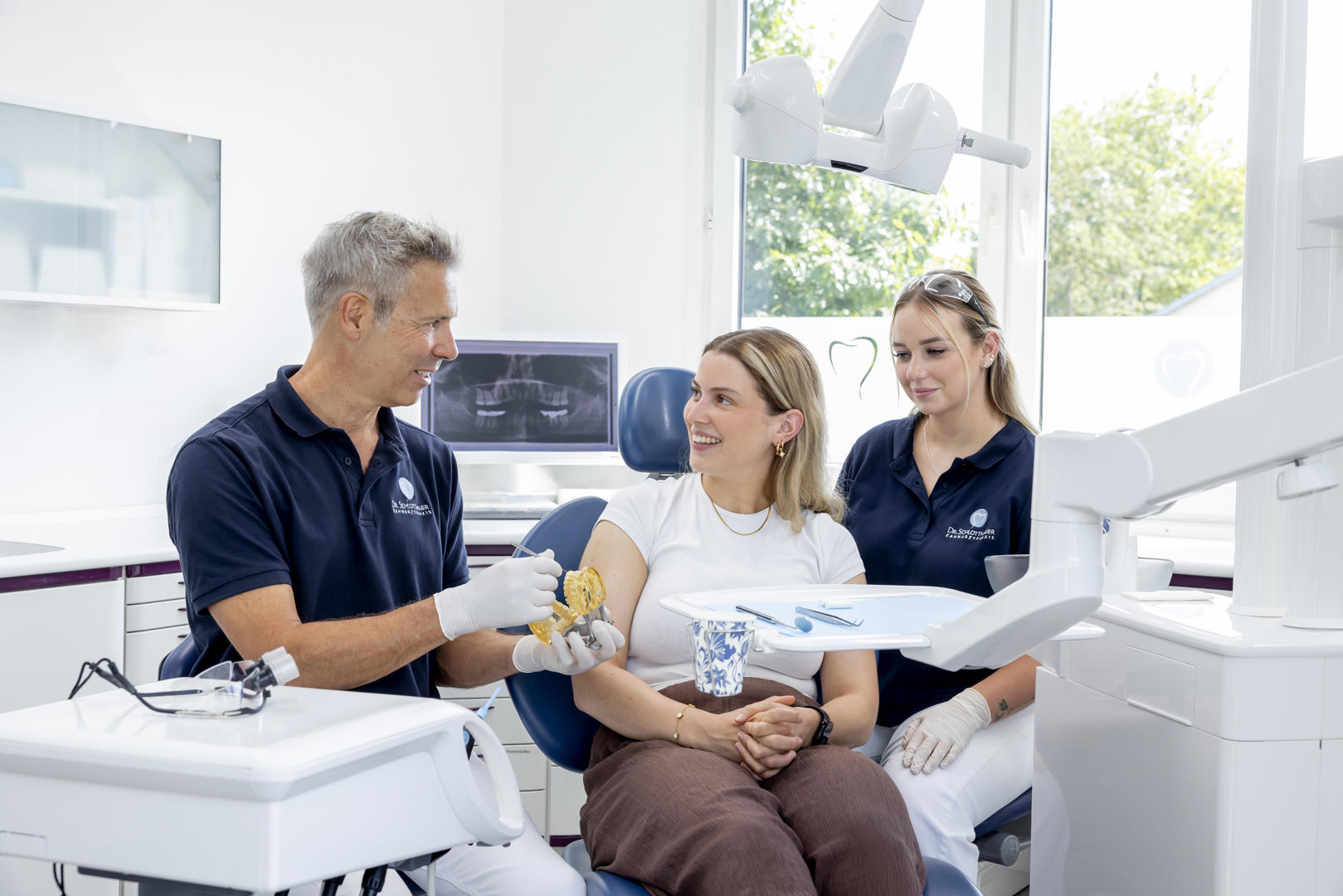 Dr. Schlotthauer erklärt einer Patientin etwas anhand eines Gebisses in der Hand, eine Mitarbeiterin schaut zu.